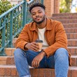 Young South African man with styled hair and beard wearing an orange jacket and jeans, holding a takeaway coffee cup on